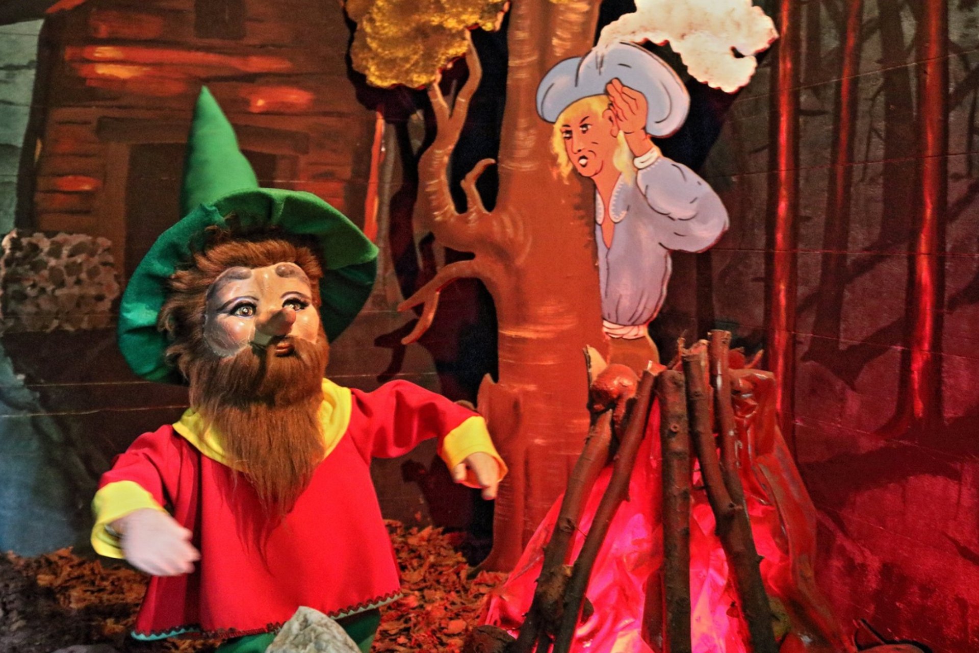 Rumpelstilzchen tanzt um das Feuer, hinter dem Baum beobachtet ihn der Bote